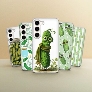 May include: Five smartphone cases featuring various pickle-themed designs. One case shows a jar of pickles, another has a pattern of pickles and dill, a central case depicts a cartoon pickle wearing a scarf, and others show pickles with sunglasses.