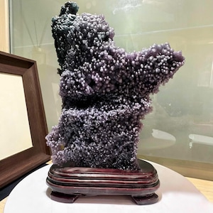 May include: A unique, natural amethyst geode specimen. The geode features a clustered, bubbly texture with shades of purple and gray. It is mounted on a dark wooden base, showcasing its organic form. The geode is a decorative item.