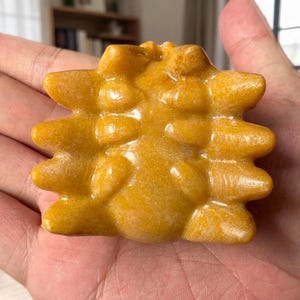 May include: A polished, golden-yellow stone carving shaped like a spider with a rounded central body and eight distinct, pointed legs extending from the sides. The surface of the mineral is smooth, glossy, and features subtle variations in color.