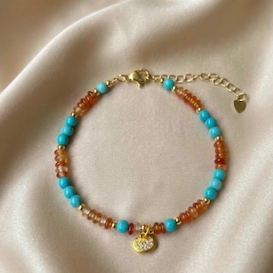 Può includere: Un braccialetto dorato con perline turchesi e ambrate. Il braccialetto ha un piccolo ciondolo dorato con chiusura a forma di cuore e un piccolo ciondolo a forma di cuore alla fine della catena regolabile. Lunghezza di circa 18 cm.