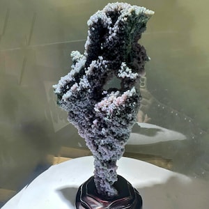 May include: A striking geode specimen with a textured, bubbly surface. The geode displays a mix of purple, grey, and pink tones, mounted on a dark wooden base. The overall shape is organic and sculptural, suitable for home decor.