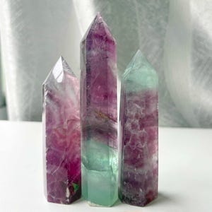 Könnte beinhalten: Drei polierte Fluorit-Kristalltürme, die aufrecht stehen. Die Kristalle haben eine hexagonale Form mit spitzen Enden und zeigen einen natürlichen Farbverlauf aus tiefem Violett, durchscheinendem Weiß und sanften Grüntönen im gesamten Stein.