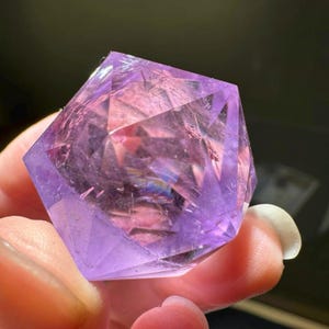 May include: A faceted, geometric amethyst crystal. The crystal is a deep purple color with internal facets that catch the light, creating a shimmering effect. The crystal is held in a person's hand.