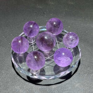 May include: A collection of seven polished amethyst spheres arranged on a circular, clear, faceted glass display stand. The purple gemstones vary slightly in size and transparency, each exhibiting natural internal inclusions and light reflections.