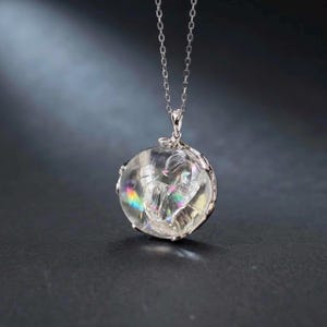 May include: A silver chain necklace featuring a round, clear, faceted pendant. The pendant contains internal fractures that create a prismatic, rainbow-colored light effect within the transparent material. The setting is a simple silver metal frame.