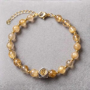 May include: Gold-toned bracelet featuring golden-yellow, translucent beads. The bracelet includes a silver-colored, geometric charm with a gold-colored design. The clasp and connecting chain are gold-colored. The bracelet is displayed on a gray surface.