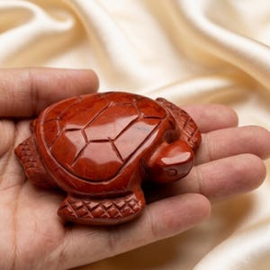 Puede incluir: Una figura de tortuga tallada en jaspe rojo. La tortuga tiene patrones detallados en el caparazón y se sostiene en una mano abierta. La figura es de color marrón rojizo y parece pulida.