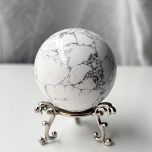 Op de afbeelding: Een witte marmeren bol met grijze adering, gepresenteerd op een decoratieve zilverkleurige standaard. Het oppervlak van de bol heeft een gebarsten patroon. De standaard heeft een ronde bovenkant en sierlijke, gebogen poten.