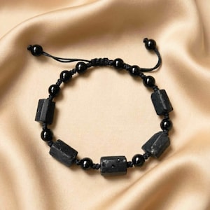 Könnte beinhalten: Schwarzes Perlenarmband mit rechteckigen und runden Perlen. Das Armband besteht aus schwarzen Perlen und ist mit einem Schiebeknotenverschluss verstellbar. Das Armband wird auf einer satinartigen Oberfläche präsentiert.