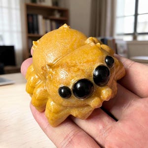 May include: A carved yellow stone figurine shaped like a jumping spider with a textured, mottled surface and four prominent, smooth, round black eyes set into the front. The object has a rounded body with small, segmented leg-like protrusions.
