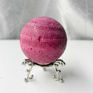 Könnte beinhalten: Eine magentafarbene Kugel mit strukturierter Oberfläche, präsentiert auf einem dekorativen silbernen Ständer. Die Kugel hat eine tiefe, satte Farbe. Der Ständer hat drei verzierte Beine und eine kreisförmige Oberseite.