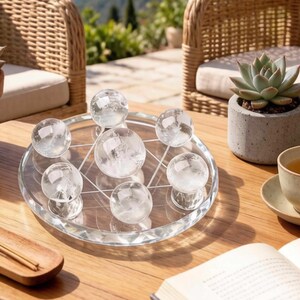 May include: A clear glass tray with a geometric design holds seven clear crystal spheres. The spheres are arranged in a pattern, with one in the center and six surrounding it. The tray sits on a wooden table.