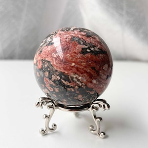 May include: A polished sphere of red and black stone, displayed on a decorative silver-coloured stand. The orb's surface features a marbled pattern of deep red and black hues. The stand has an ornate, three-legged design.
