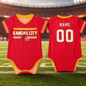 May include: A red baby bodysuit with yellow trim, featuring the text "KANSAS CITY" and a football helmet graphic on the front. The back displays the word "NAME" above the number "00" in white with yellow outlines, mimicking a sports jersey style.