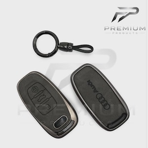 May include: Black Audi car key fob covers and a key ring. The key fobs are dark grey with the Audi logo and key buttons. The key ring is black with a small strap.