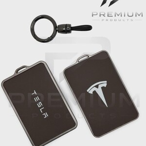 May include: Two black Tesla key fob holders with silver trim, one showing the Tesla logo and the other the word "TESLA". A black key ring with a leather strap is also shown. The "Premium Products" logo is in the upper right corner.
