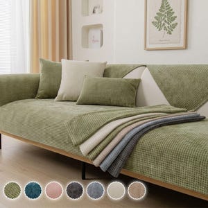 May include: A sage green sofa cover with a waffle-textured pattern, featuring a matching square pillow, a rectangular pillow, and a draped throw. The cover is displayed on a sofa, with seven circular colour swatches shown at the bottom.