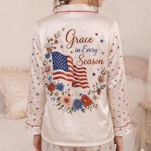 May include: Cream-coloured pyjama set with an American flag design and floral wreath. The top's back displays the text "Grace in Every Season". The set includes a long-sleeved top and matching trousers.