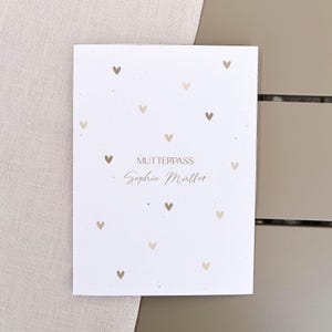 Könnte beinhalten: Ein weißes, rechteckiges Heftcover mit einem Muster aus kleinen beigen Herzen, die über die Oberfläche verteilt sind. Der Text MUTTERPASS ist in einer einfachen Schrift über dem Namen Sophie Müller gedruckt, der in beiger Schreibschrift steht.