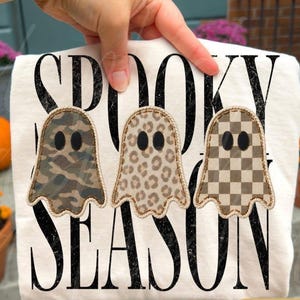 May include: A white t-shirt featuring the black distressed text SPOOKY SEASON. Three ghost-shaped patches are arranged horizontally across the centre. The first ghost has a camouflage pattern, the second a leopard print, and the third a checkerboard.