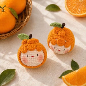 May include: Two handmade crochet orange-shaped toys with smiling faces, green leaves, and brown stems. The toys are orange and white, with pink cheeks and small red mouths. Fresh oranges and a basket are in the background.