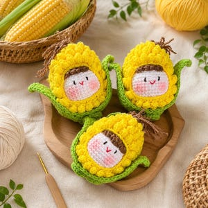 May include: Three crocheted corn-shaped dolls with yellow kernels, green husks, and brown hair. Each doll features a white face with black eyes, a red v-shaped mouth, and pink cheeks. The dolls are arranged on a wooden tray with a crochet hook nearby.