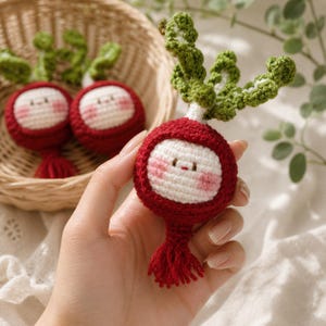 May include: Crocheted radish toy with a smiling face, red body, white face, and green leafy top. The radish has a red tassel at the bottom. Two more radishes are in a wicker basket in the background.