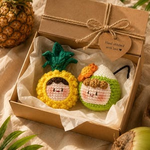 May include: A gift box contains two crocheted tropical fruit ornaments: a yellow pineapple and a green coconut, each with a smiling face. The box is tied with twine and a heart-shaped tag that reads "Hand-stitched with love".