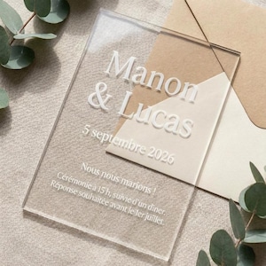 May include: A clear rectangular acrylic wedding invitation featuring white printed text that reads Manon & Lucas, 5 septembre 2026, and additional event details, placed over a tan envelope with a pointed flap and accented by green eucalyptus leaves.