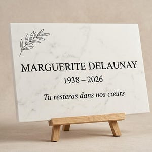 Puede incluir: Una placa rectangular de mármol blanco con vetas grises, expuesta sobre un pequeño caballete de madera. La placa presenta un dibujo lineal negro de una rama de olivo, el nombre MARGUERITE DELAUNAY, las fechas 1938-2026 y el texto Tu resteras dans nos cœurs.