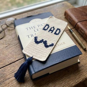 May include: A rectangular cream-colored crocheted bookmark featuring the dark blue stitched text DAD and a dark blue mustache graphic. The bookmark is finished with a dark blue yarn tassel attached to the bottom edge, creating a textured design.
