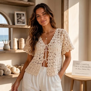 May include: Cream-colored, open-front crochet cardigan with short sleeves. The cardigan has a scalloped edge and a tie closure. The text on the sign reads "THE COASTAL LACE CARDIGAN - An Artisan Collection Piece". The cardigan is part of a coastal collection.
