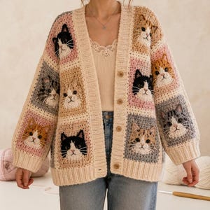 Peut inclure: Cardigan en crochet de couleur crème avec des carrés représentant des visages de chats de différentes couleurs. Le cardigan a une fermeture boutonnée sur le devant et des manches longues. Le texte "Cat Cardigan Pattern PDF" est affiché en bas de l'image.