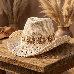 Può includere: Un cappello da cowboy all'uncinetto color crema con una tesa larga e una fascia decorativa di fiori marroni. Il cappello è appoggiato su un tavolo di legno rustico, con piante secche sullo sfondo. La tesa del cappello ha un design traforato delicato.