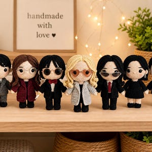 May include: A collection of seven handmade crochet dolls, each with unique hairstyles, outfits, and accessories. The dolls are holding hands and standing on a wooden shelf. A framed sign behind them reads "handmade with love".