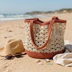 Pode incluir: Uma sacola tote creme, com tecido aberto, com acabamentos e alças cor de ferrugem está em uma praia arenosa ao lado de um chapéu de palha e óculos de sol. A sacola está cheia de itens, sugerindo um dia na praia.