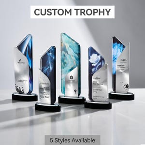 May include: Five custom trophies with unique designs. Each trophy features a clear, angled glass front with various artistic backgrounds and a silver metal backing. The bases are black and circular. Text on the trophies includes "CUSTOM TROPHY" and "5 Styles Available."