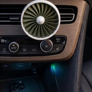 May include: A white and olive green car air vent fan is mounted on a car's dashboard. The fan has a circular design with angled blades. The dashboard has wooden accents and control buttons for climate and other car functions. A smartphone rests in a compartment below.