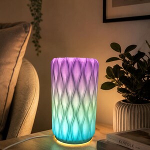 Puede incluir: Lámpara cilíndrica con un diseño ondulado y texturizado, iluminada con un degradado de colores de azul a verde a morado. La lámpara está sobre una superficie de madera junto a libros y una planta en maceta.