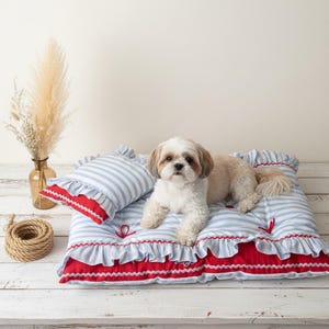 Puede incluir: Un perro pequeño descansa sobre una cama para mascotas a rayas azules y blancas con una almohada a juego. La cama y la almohada tienen bordes rojos con volantes. Un jarrón con flores secas y una bobina de cuerda están en el fondo.