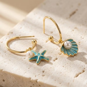 Könnte beinhalten: Ein Paar goldfarbene Creolen. Ein Ohrring hat einen blauen Seestern-Anhänger mit einer weißen Perle, der andere einen blauen Muschel-Anhänger mit einer weißen Perle. Die Ohrringe sind vor einem neutralen Hintergrund platziert.