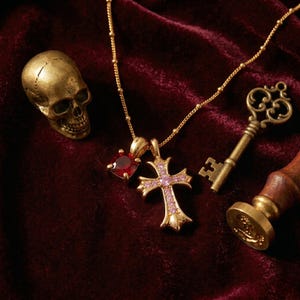 Può includere: Un teschio color oro, un ciondolo con rubino e un ciondolo a croce con pietre preziose rosa sono esposti su una superficie di velluto bordeaux. Sono presenti anche una chiave d'oro e un sigillo di cera, creando un'estetica gotica o vintage.