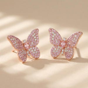 May include: A pair of rose gold butterfly stud earrings. Each earring features a butterfly design with pink and white gemstones. The earrings are set against a soft, neutral background, highlighting their delicate details.