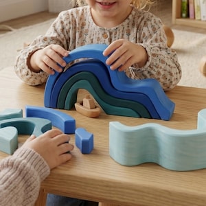 Peut inclure: Un ensemble de jouets en bois comprenant un empileur arc-en-ciel en forme de vague bleu et vert, un petit bateau en bois et d'autres blocs bleus. Le jouet est conçu pour les enfants et est présenté sur une table en bois de couleur claire.