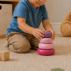 Peut inclure: Un enfant empile un ensemble de quatre anneaux en bois de couleurs violettes et roses. Les anneaux sont de tailles graduées, avec la couleur la plus foncée à la base. L'enfant porte un t-shirt bleu et un pantalon kaki.