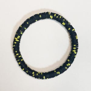 Peut inclure: Un bracelet de perles noires et jaunes. Le bracelet est composé de petites perles rondes et plates. Les perles sont principalement noires avec de petits accents jaunes. Le bracelet est circulaire et repose sur une surface blanche.