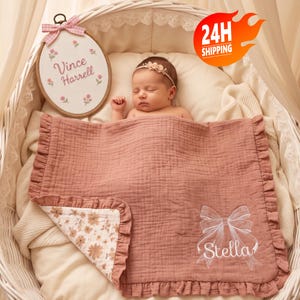Könnte beinhalten: Eine altrosa Musselin-Babydecke mit gerüschten Rändern, verziert mit einer weißen Stickerei in Schleifenform und dem Namen Stella. Die Decke hat ein florales Innenfutter und liegt in einem weißen Stubenwagen mit einem ovalen Schild Vince Harrell.