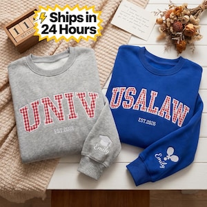 May include: Two sweatshirts, one gray and one blue, are displayed. The gray sweatshirt has "UNIV" in red gingham lettering, and the blue one says "USALAW." Both have "EST.2026" and personalized sleeve details. The image also includes a wooden calendar.