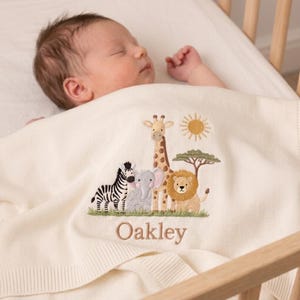 Könnte beinhalten: Eine cremefarbene Strickdecke für Babys mit einer Stickerei, die ein Zebra, einen Elefanten, eine Giraffe und einen Löwen im Gras unter einer Sonne zeigt. Der Name Oakley ist in braunem Garn unter den Tieren auf dem weichen Stoff eingestickt.