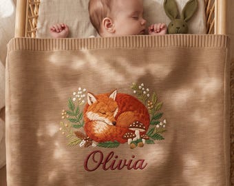 May include: A beige knitted baby blanket featuring an embroidered design of a sleeping fox surrounded by green foliage and small mushrooms. The name Olivia is embroidered in dark red script below the fox. The blanket has a ribbed top border.